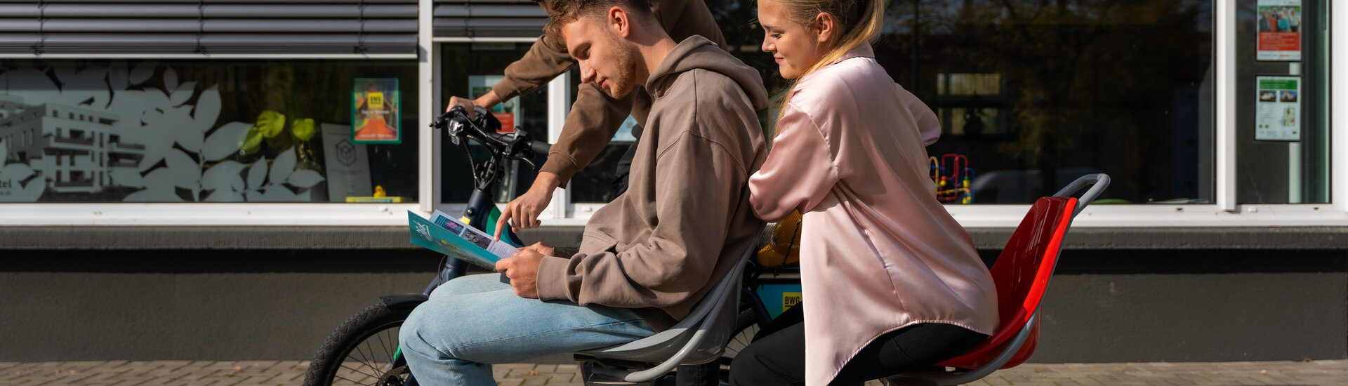 Zwei Personen auf hintereinander stehenden Stühlen lesen den Blick, im Hintergrund ist eine Person auf einem Fahrrad, die auf den Blick zeigt